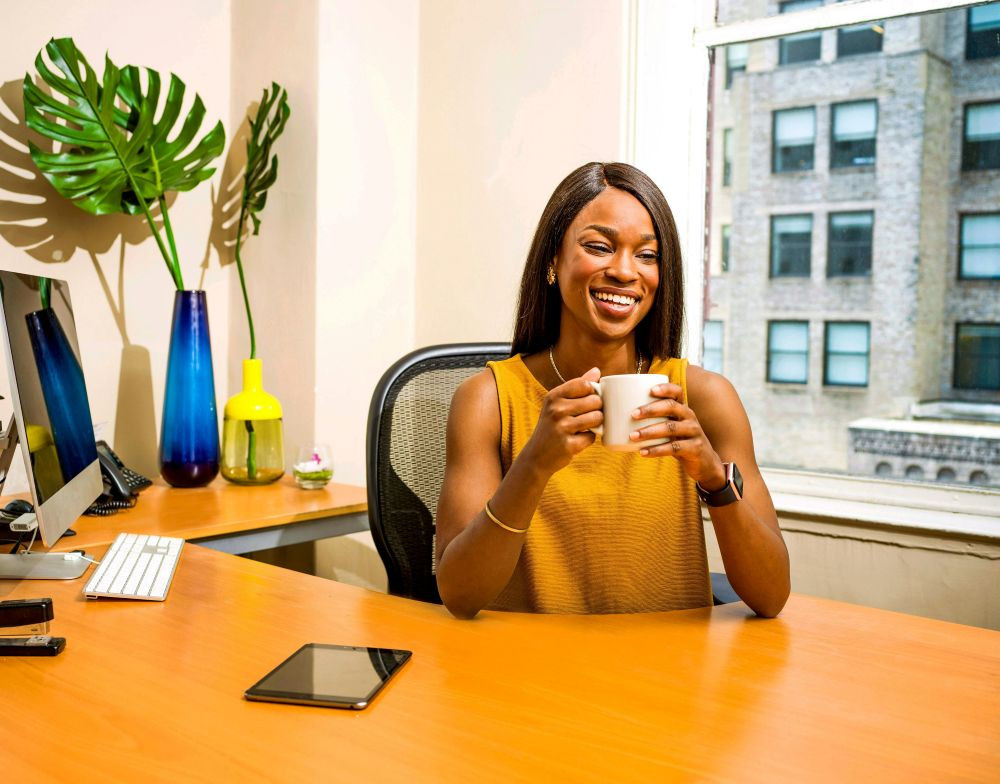 An administrative professional smiling at her desk with a coffee mug in a sunlit office